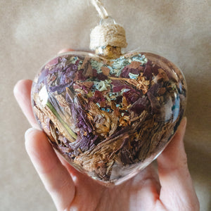 Hand holding a glass heart ornament filled with rose gold lilies, red rose petals and blue hydrangeas.