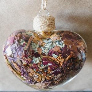 Top view of a glass heart filled with rose gold lilies, red rose petals and blue hydrangeas.